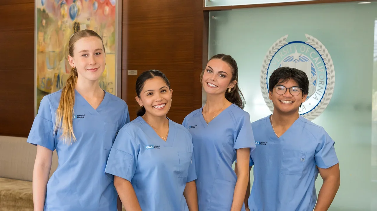 4 nursing students wearing blue scrubs smiling for a photograph in the Factor Building lobby with the new Joe C. Wen School of Nursing sign visible.