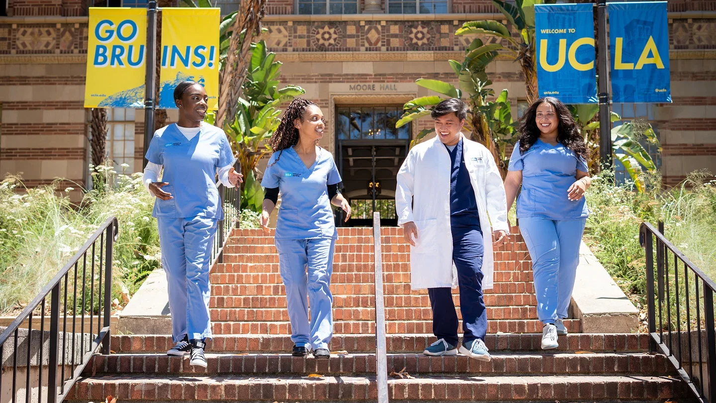 Five people wearing light blue medical scrubs and one white lab coat walk down brick steps on a university campus. Banners on lampposts read “GO BRUINS!” and “WELCOME TO UCLA."