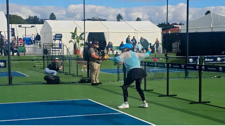 A woman playing pickleball wearing a white and blue jacket, black skirt, black leggings, and blue hat.