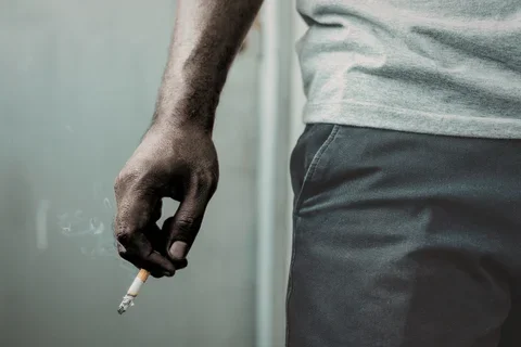 Close-up of a person’s hand holding a lit cigarette with smoke rising, next to their hip, wearing a light gray shirt and dark pants against a neutral background.