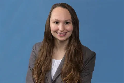 Katie Kozakowski in a grey jacket and white shirt against a blue backdrop.