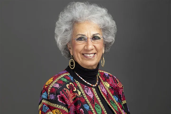 Afaf Meilis is shown in a studio portrait against a gray background, wearing glasses, hoop earrings, a beaded necklace, and a multicolored patterned jacket over a dark top. 
