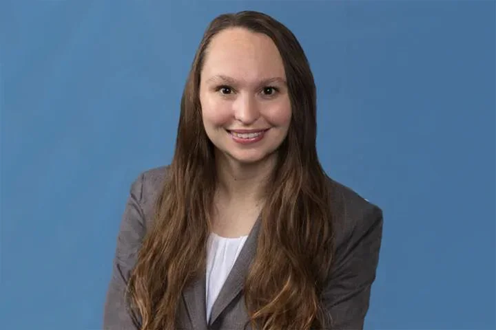 Katie Kozakowski in a grey jacket and white shirt against a blue backdrop.