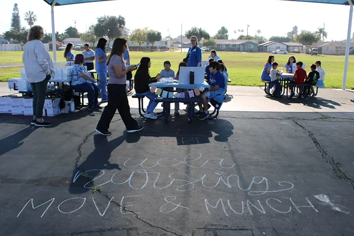 Children and UCLA Nursing students sitting on playground tables with the words Move and Munch written in chalk on the ground.