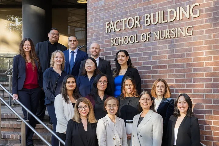 Fifteen DNP students, class of 2026, standing staggered in rows outside on the front steps of the School of Nursing building. Students are dressed in professional clothing and are smiling.
