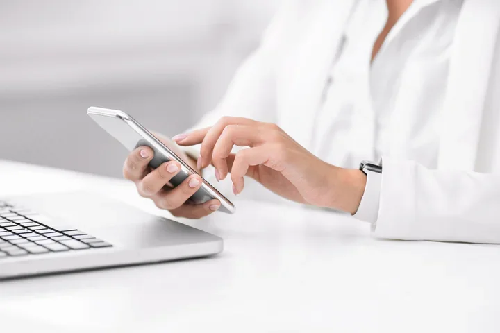 Close-up of a person’s hands using a smartphone on a white desk, with an open laptop nearby; the person wears a white long-sleeve shirt and a smartwatch, in a bright, minimal workspace.