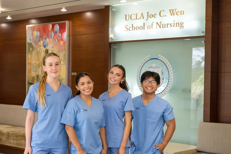 4 nursing students wearing blue scrubs smiling for a photograph in the Factor Building lobby with the new Joe C. Wen School of Nursing sign visible.