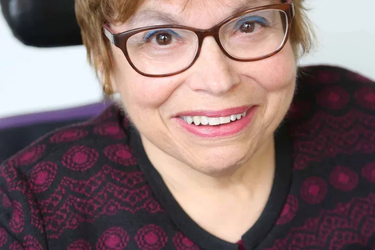 A headshot of Judy Heumann, a cis-gender white woman who is a wheelchair user with short brown hair. She is wearing brown glasses, a maroon and black embroidered cardigan with the top buttoned, and a matching maroon shirt. She is smiling kindly. 