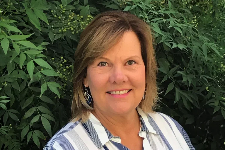 Karen Nelson wearing a white and blue stripped shirt standing in front of greenery. 