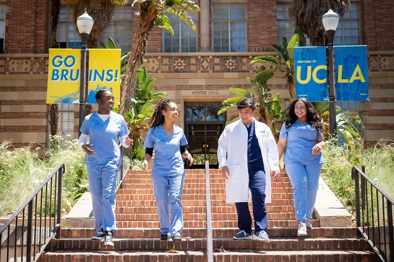 Five people wearing light blue medical scrubs and one white lab coat walk down brick steps on a university campus. Banners on lampposts read “GO BRUINS!” and “WELCOME TO UCLA."