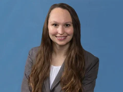 Katie Kozakowski in a grey jacket and white shirt against a blue backdrop.