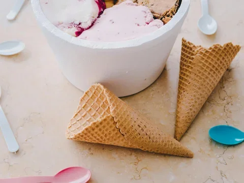Tub of assorted ice cream flavors with waffle cones and colorful spoons on a table.
