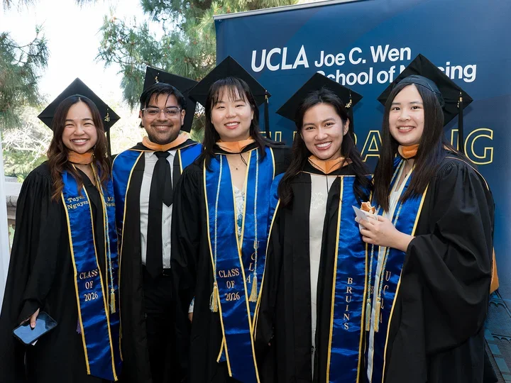 5 UCLA nursing students wearing their graduation caps and gowns standing in front of a UCLA Joe C. Wen School of Nursing sign.