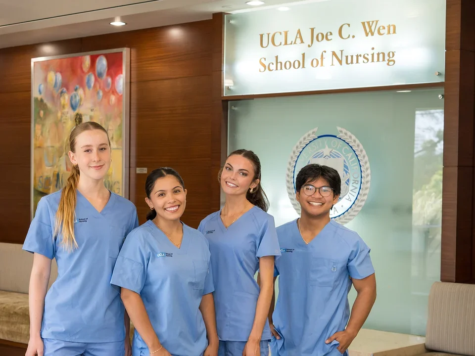 4 nursing students wearing blue scrubs smiling for a photograph in the Factor Building lobby with the new Joe C. Wen School of Nursing sign visible.