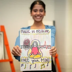 A student holding up their art project, a painting with the words "music is medicine"