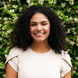 Alexis Ross wearing a tan dress standing in front of greenery. 