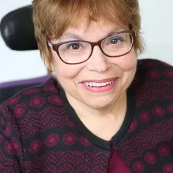 A headshot of Judy Heumann, a cis-gender white woman who is a wheelchair user with short brown hair. She is wearing brown glasses, a maroon and black embroidered cardigan with the top buttoned, and a matching maroon shirt. She is smiling kindly. 