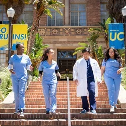  Five people wearing light blue medical scrubs and one white lab coat walk down brick steps on a university campus. Banners on lampposts read “GO BRUINS!” and “WELCOME TO UCLA,” with a brick academic building, palm trees, and landscaping in the background.