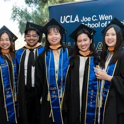 5 UCLA nursing students wearing their graduation caps and gowns standing in front of a UCLA Joe C. Wen School of Nursing sign.