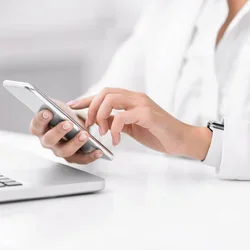 Close-up of a person’s hands using a smartphone on a white desk, with an open laptop nearby; the person wears a white long-sleeve shirt and a smartwatch, in a bright, minimal workspace.