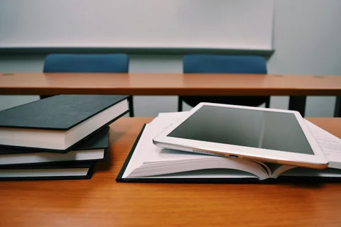 a classroom desk with books stacked on the left, a book open on the right with a tablet on top and an empty desk in the back with two blue chairs in front of a whiteboard